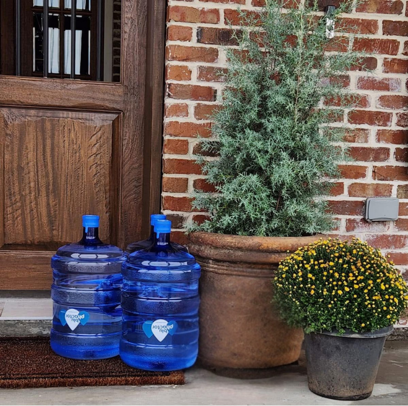 Two Blu Phactor water delivery jugs by a front door with potted plants.